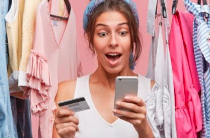 Mulher surpresa fazendo compras de roupas em loja, segurando cartão de crédito e celular, com roupas coloridas ao fundo, explorando ofertas de moda.