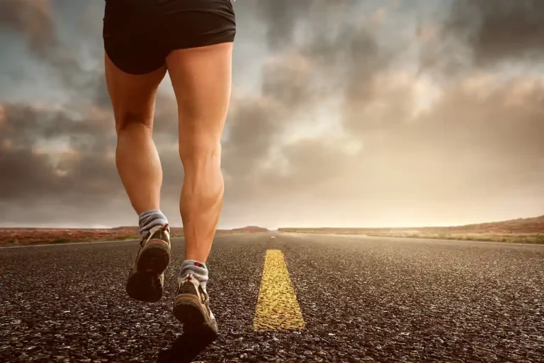 Imagem de uma pessoa correndo na pista, focada nas pernas musculosas, em uma estrada deserta sob céu nublado, representando corrida e fitness.
