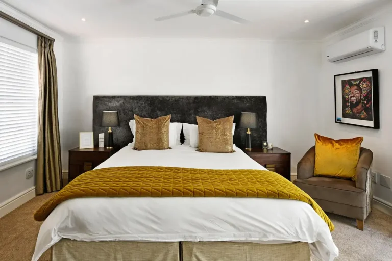Quarto moderno e aconchegante com cama king size, almofadas douradas, poltrona confortável com almofada amarela, iluminação suave e decoração elegante.